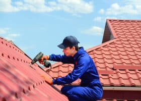 Close up of young happy man contractor worker in blue overalls is repairing a red roof with electric screw driver. He is smiling. Roofing concept.